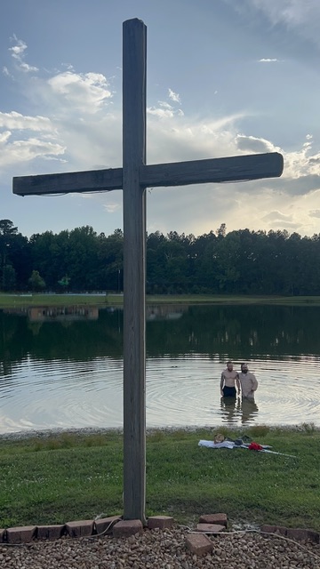 A tall cross beside the lake with a baptism happening in the water